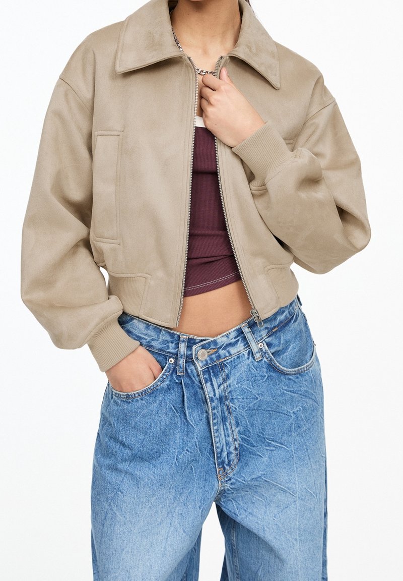Personne portant une veste beige zippée sur un crop top bordeaux et un jean en denim bleu taille haute, une main dans la poche.