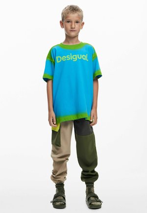 Plavokosi dječak stoji, nosi plavu i zelenu Desigual majicu, hlače od zakrpa u bež i zelenoj boji, zelene čarape i crne sandale.