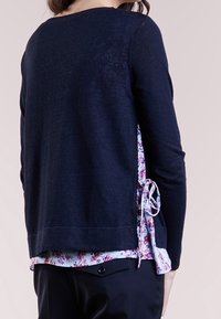 Mörk marineblå tröja med rund halsringning och långa ärmar, lagd över en ljusblommig skjorta med knytning på sidan.