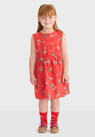 Smilende pige med rødt hår iført en ærmeløs rød kjole og matchende sokker med ko- og kaktusprint samt brune sko.