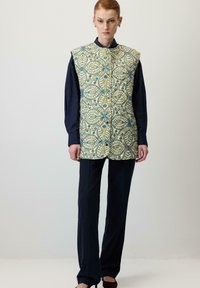 Gequilted vest in een geel en blauw bloemenpatroon, met een hoge kraag, vooraan knopen, gedragen over een donker langslijfs T-shirt en een broek.