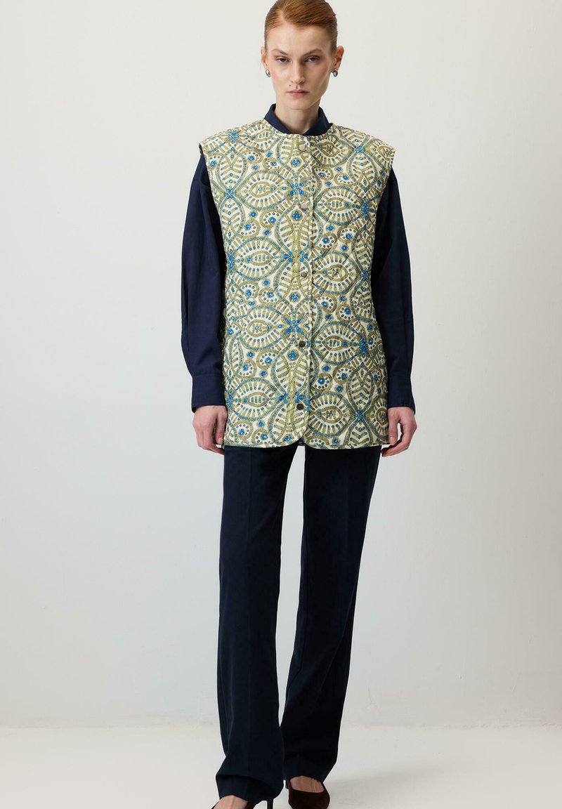 Gequilted vest in een geel en blauw bloemenpatroon, met een hoge kraag, vooraan knopen, gedragen over een donker langslijfs T-shirt en een broek.