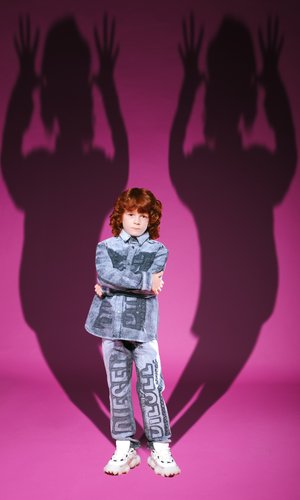 Denim-Outfit in blaugrau mit großen schwarzen "DIESEL"-Buchstaben. Das Kind posiert vor einem pinken Hintergrund und wirft lange Schatten an die Wand.