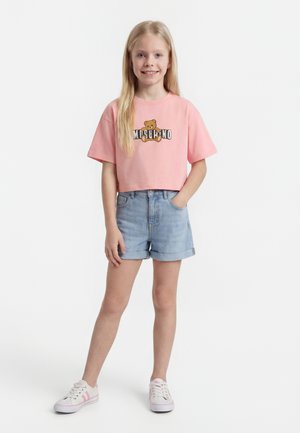 Jauna meitene valkā rozā apgrieztu topu ar lācēna grafiku, džinsa šortus un baltus sporta apavus, stāv ar vienu roku kabatā.