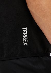 Tissu polaire noir avec logo brodé "TERREX" blanc et vue partielle du bras d’une personne portant le vêtement.