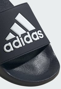 Sandale noire à glissière en matériau synthétique texturé, avec un logo Adidas blanc bien visible sur la sangle et une semelle à motifs.