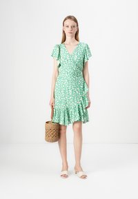 Grønn omslagskjole med hvitt abstrakt mønster, korte volangerdetaljer, og tiered hem. Kombinert med en vevd veske og lette sandaler.