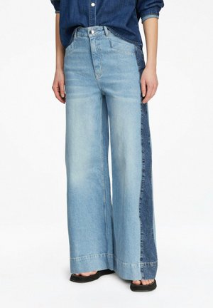 Kobieta w wysokich, szerokich spodniach jeansowych z panelami z jasnego i ciemnego denimu, w połączeniu z czarnymi sandałami z odkrytymi palcami i podwiniętą jeansową koszulą.