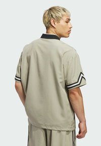 Béžová košeľa s krátkym rukávom a čiernym golierom, s manžetami v chevronovom vzore. Hladká tkanina s voľným strihom, obsahuje čierne akcentové pruhy.