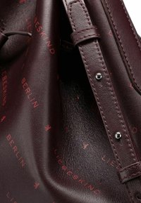 Wnętrze burgundowej skórzanej torebki z wytłoczonym wzorem logo w kontrastowej czerwieni. Gładka tekstura, regulowany pasek i srebrne dodatki.