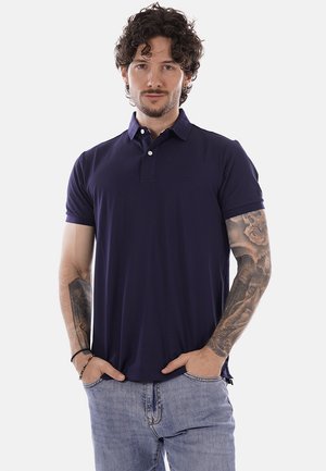 Uomo con capelli ricci e tatuaggi su entrambe le braccia, indossa una polo blu navy e jeans azzurri, con le mani nelle tasche, rivolto in avanti.