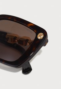 Motoškrupinové slnečné okuliare s hnedými sklami, zlatými akcentmi a logom Swarovski na skle. Textúrovaný rám a hrubé nožičky.