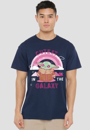 Junger Mann trägt ein marineblaues T-Shirt mit einer grünen Alien-Cartoon-Figur unter einem pinken Regenbogen und dem Text „Süßester in der Galaxie."