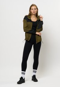 Giubbotto leggero verde oliva e nero con cappuccio, chiusura con zip e polsini elastici, abbinato a leggings neri aderenti e sneakers nere.
