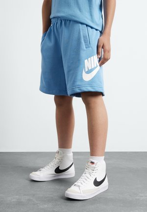 Person iført lyseblå Nike-shorts og hvide højtstående Nike-sneakers, der står på et gråt gulv mod en ensfarvet baggrund.