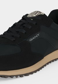 Baskets GANT noires et beiges avec lacets noirs, semelle texturée et logo sur le côté et la languette, vues de côté et de face.