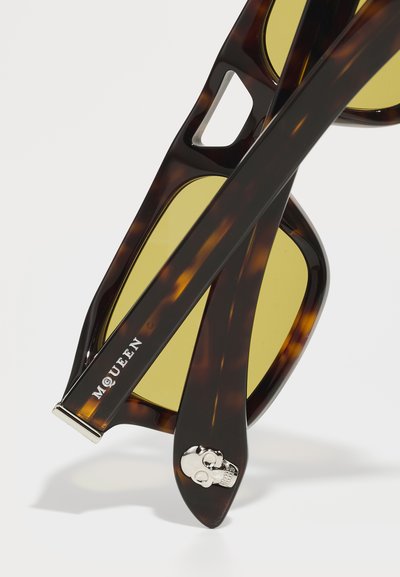 Ochelari de soare cu mototol, cu lentile galbene, având un accent de craniu argintiu pe lateral și un design distinctiv tăiat în montură.