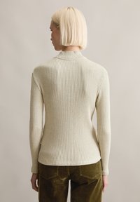 Rippiges, cremefarbenes Langarmoberteil mit hohem Kragen, strukturiertem Stoff, taillierter Form und abgerundetem Saum, kombiniert mit dunkelgrünen Cordhosen.