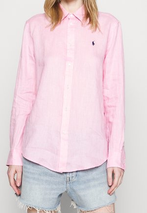 Personne portant une chemise à manches longues rose clair à boutons avec un petit logo foncé et un short en jean délavé bleu clair effiloché.