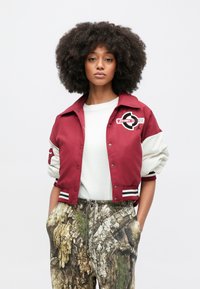 Κόκκινο και λευκό κοντό μπουφάν με λογότυπο στο στήθος, συνδυασμένο με παντελόνι σε καμουφλάζ σχέδιο και λευκό πουκάμισο με στρογγυλή λαιμόκοψη.