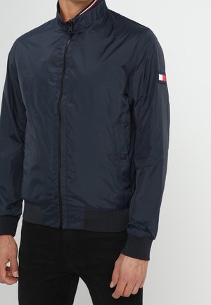 Mand iført en navyblå bomberjakke med lynlås, ribstrikkede manchetter og kant, lille logopatch på venstre ærme.