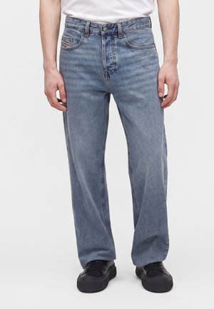 Homme portant un jean droit bleu clair et des baskets noires, debout devant un fond clair uni.