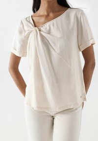 Femme portant un top à manches courtes blanc cassé avec un détail de nœud tordu au niveau de l'encolure et un pantalon blanc cassé assorti.