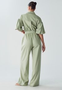 Moteris su bandeliu, apsirengusi šviesiai žaliu dirželiu per juosmenį kombinezonu su plačiomis kelnėmis ir pūkuotomis rankovėmis, stovi prieš paprastą foną.