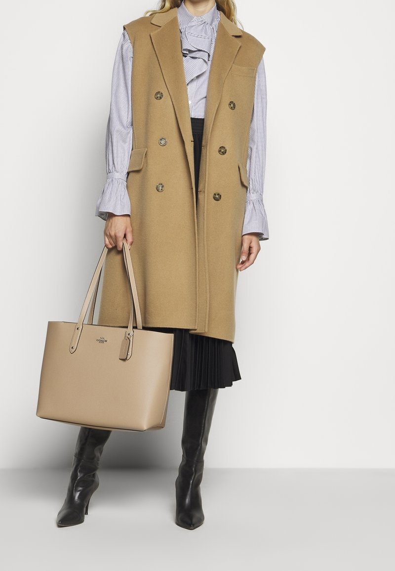 Femme portant un manteau sans manches beige sur un chemisier à volants et une jupe plissée, tenant un grand sac cabas beige, portant des bottes noires jusqu’au genou.