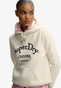 Crème hoodie met een trekkoord en een voorzak, met zwarte tekst "SuperDry" en "J'ADORE PARIS". Gelaagd over een roze gestreepte shirt.