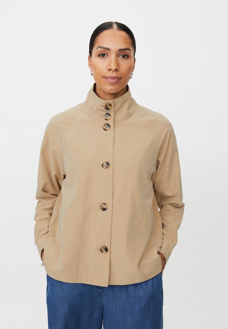 Béžová bunda s vysokým límcem, zapínáním na knoflíky vpředu a raglánovými rukávy; hladká struktura látky; vybavena velkými, průhlednými knoflíky.