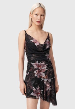 Femme aux longs cheveux blonds portant une robe noire sans manches à fleurs roses et blanches avec un ourlet asymétrique.