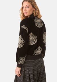 Chaleco bomber de terciopelo negro con intrincados bordados dorados en patrones circulares y florales, con puños y dobladillo acanalados.