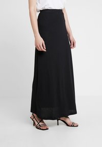Falda maxi negra con un diseño sencillo, cinturilla elástica y tela fluida que llega hasta los tobillos. Llevada con sandalias negras de tiras.