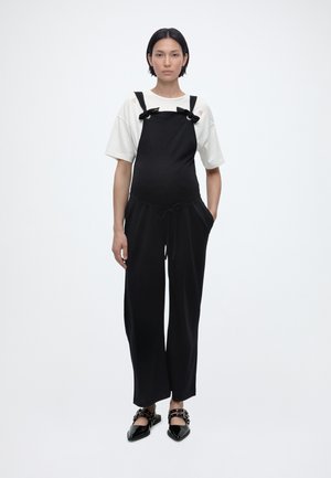 Γυναίκα που στέκεται, φοράει μαύρη ολόσωμη φόρμα με πλατιά πόδια και τιράντες με φιόγκο πάνω από λευκό κοντομάνικο πουκάμισο, και μαύρα παπούτσια μυτερά με αγκράφες.