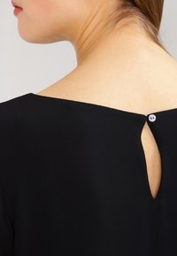 Haut noir avec un large décolleté, un petit détail œillet à l'arrière et un seul bouton décoratif au niveau du cou. Texture de tissu lisse.