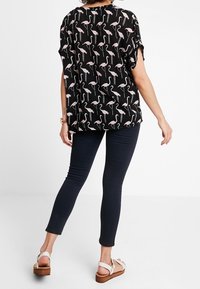 Schwarze Bluse mit pinkem Flamingo-Muster, lockere Passform, kurze Ärmel. Kombiniert mit dunkelblauen Skinny-Jeans und braunen Plateausandalen.