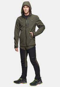 Oliwkowa kurtka softshell z kapturem, zapinana na suwak kieszeń na piersi oraz czarne spodnie z kontrastowymi panelami bocznymi i wytrzymałe buty z zielonymi akcentami.