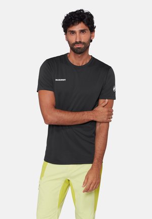 Mand med mørkt krøllet hår og skæg iført en sort Mammut T-shirt og lysegule bukser, stående med den ene arm på tværs af kroppen.