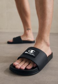 Des sandales noires à glissière en matériau synthétique lisse, dotées d'une large sangle avec un logo blanc, d'une semelle basse et d'une conception à bout ouvert.