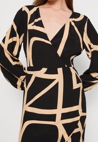 Schwarzes Wickelkleid mit langen Ärmeln und tiefem V-Ausschnitt, verziert mit einem beigen geometrischen Muster, aus einem glatten, leichten Stoff gefertigt.