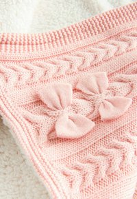 Couverture en tricot rose clair avec des nattes, des motifs surélevés et des nœuds. Finition texturée avec un dessous doux et pelucheux.