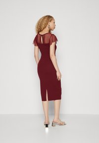 Robe midi bordeaux avec des manches flottantes transparentes, détail de dos en trou de serrure et fente sur le côté. Associée à des sandales à talons métalliques.