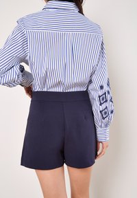 Weißes Hemd mit blauen Streifen und Puffärmeln, kombiniert mit dunkelblauen, hoch taillierten Shorts mit glatter Textur und ohne sichtbare Taschen.