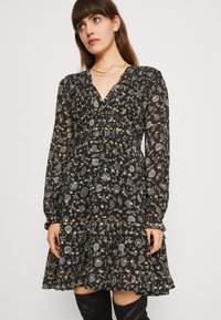 Vestido preto de mangas longas e decote em V, com um padrão floral em amarelo e branco. Apresenta uma bainha com babados e detalhe de cintura franzida.