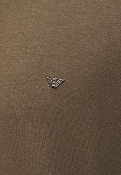 Καφέ ύφασμα με ένα μικρό, κεντρικό κεντημένο έμβλημα που μοιάζει με στυλιζαρισμένα φτερά ή πουλί σε ανοιχτό γκρι κλωστή.