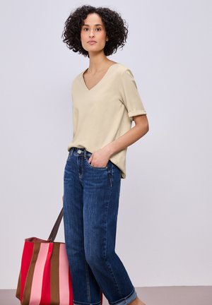 Kvinde med krøllet hår iført en beige V-hals skjorte og blå jeans, der holder en stor stribet shoppertaske med røde, pink og brune striber.