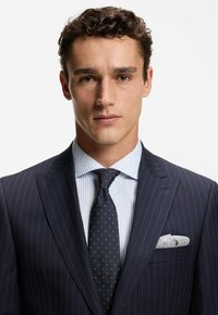 Completo a righe sottili blu navy con camicia a righe bianche e blu. Presenta una cravatta scura a pois e un fazzoletto grigio nel taschino della giacca.