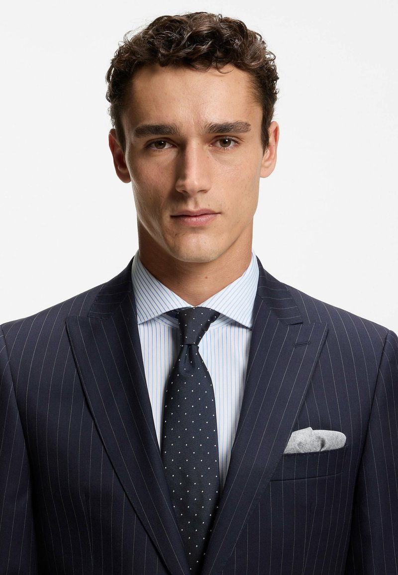 Completo a righe sottili blu navy con camicia a righe bianche e blu. Presenta una cravatta scura a pois e un fazzoletto grigio nel taschino della giacca.