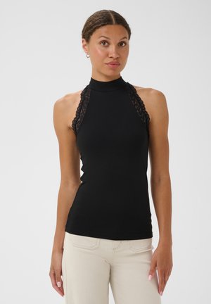 Femme portant un haut noir sans manches à col halter avec une bordure en dentelle et un pantalon couleur crème, debout devant un fond blanc uni.